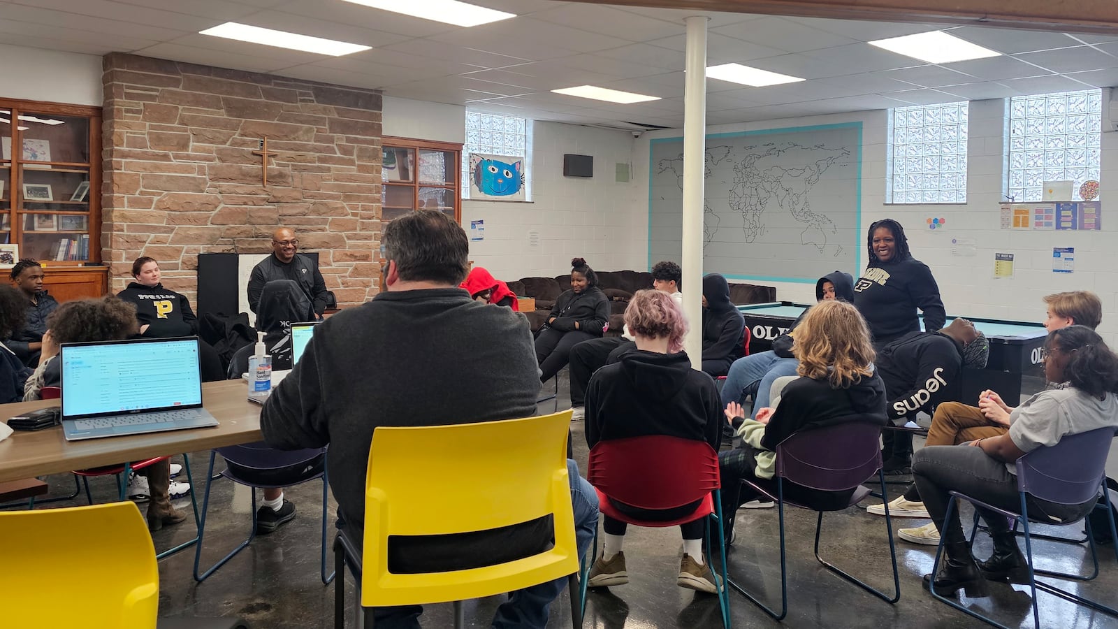 A group of adults and high school students gather around in a wonky circle in a room with a large brick wall in the back.