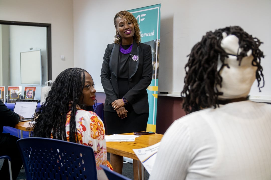 A woman wearing a dark-colored suit stands at the front of a room, smiling and looking at two women whose backs are to the camera.