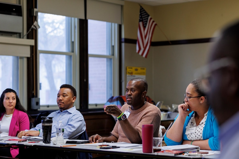 Four people sit next to each other in a row at a desk with a classroom setting and American flag in the background.