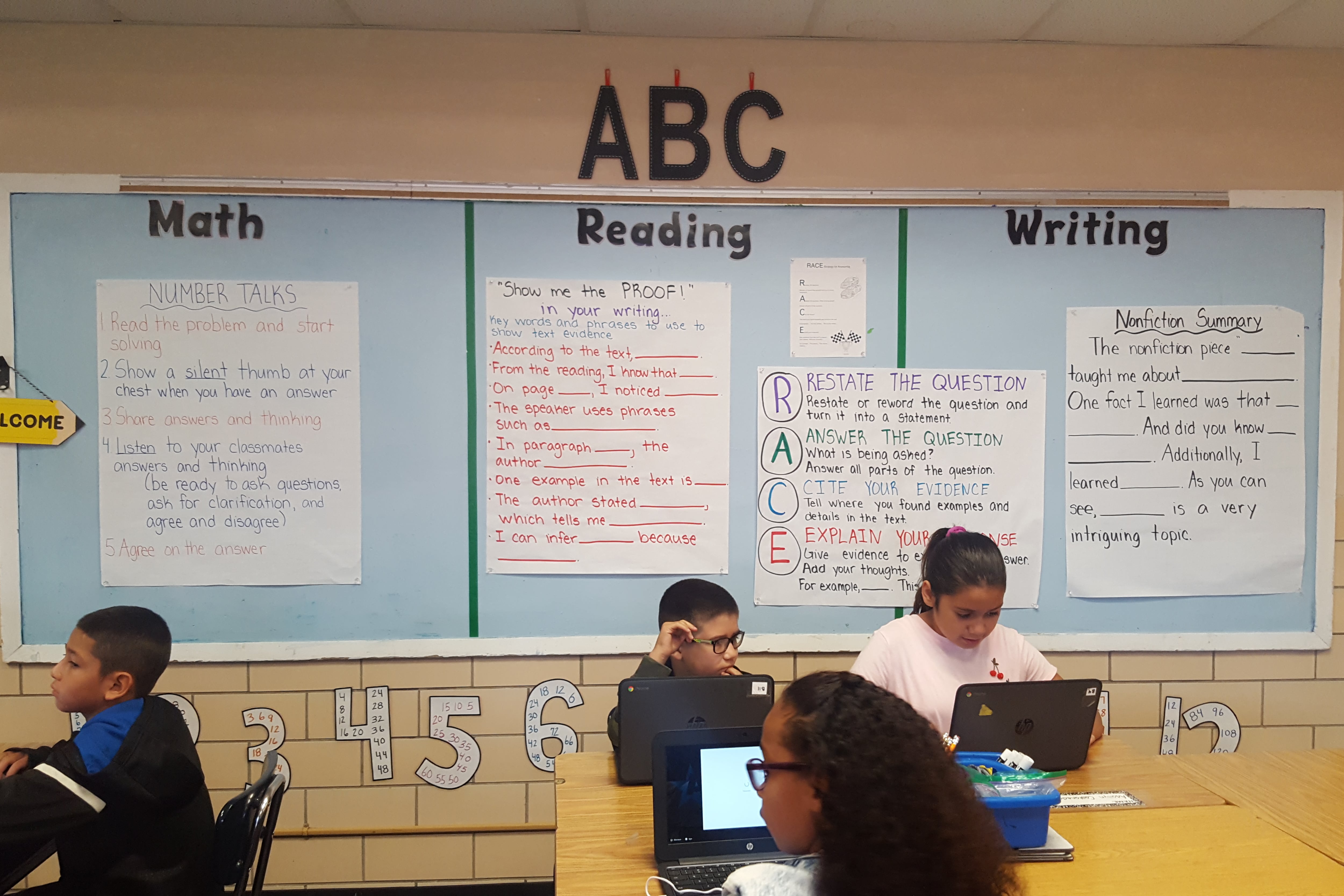 Students sit at tables in front of a bulletin board.