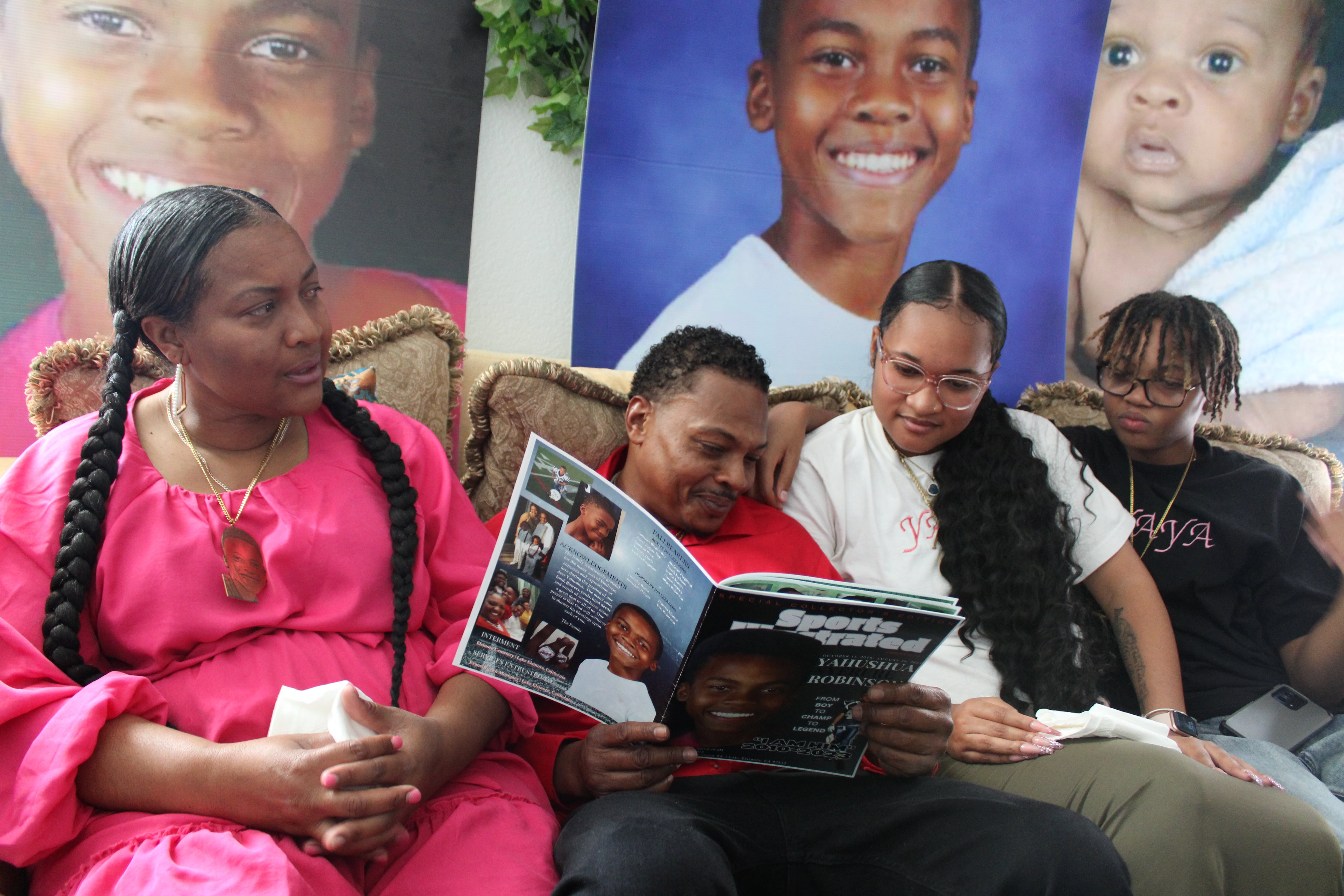 A mother, father, and two children sit on a couch looking at pictures of their brother, who died at age 12. Large photos of their brother are also on the wall behind them, looking over them.