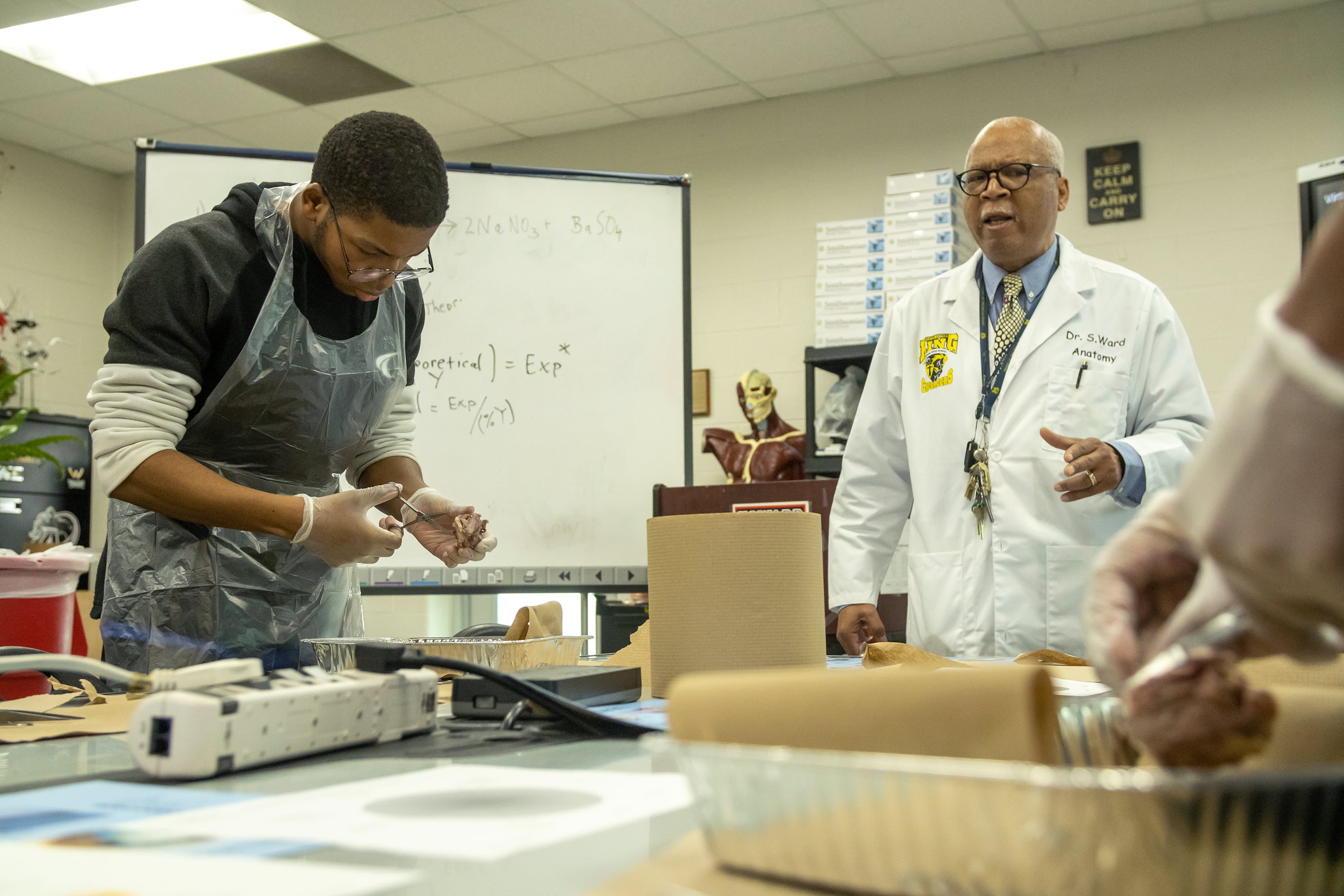 Two people wearing science lab gear stand in a classroom at a table with a whiteboard in the background.