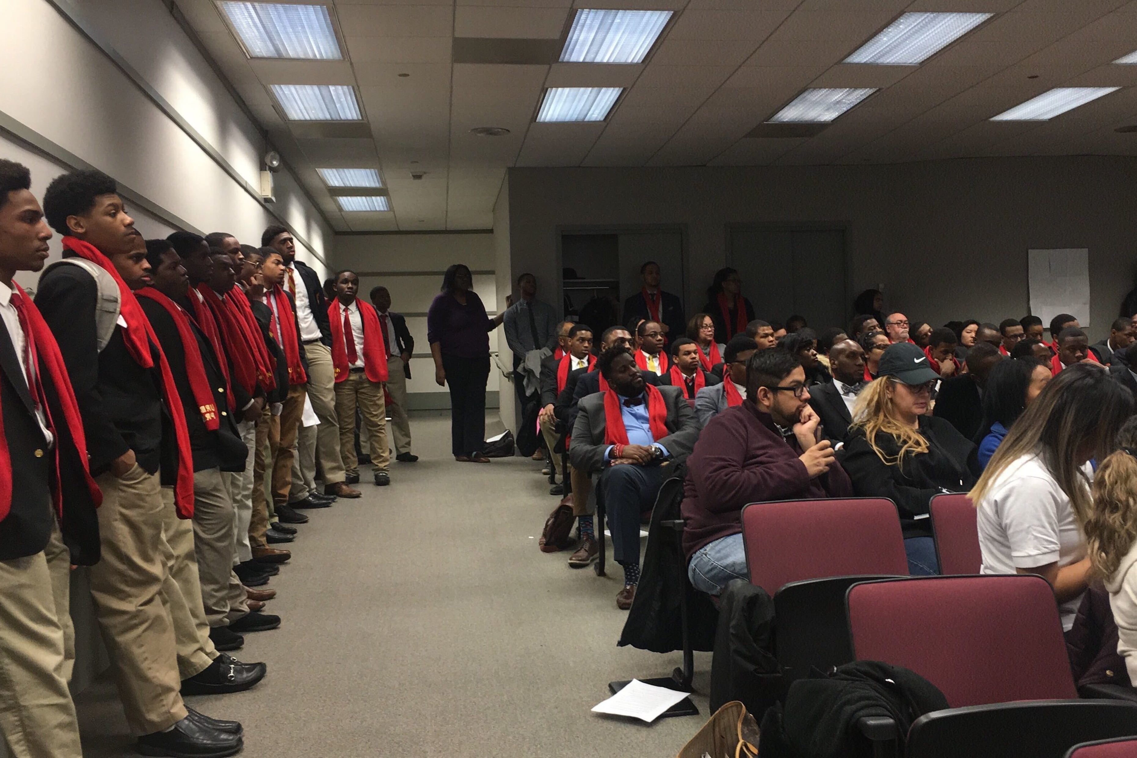 A group of high school students stand at left. Adults are seated nearby in an auditorium.