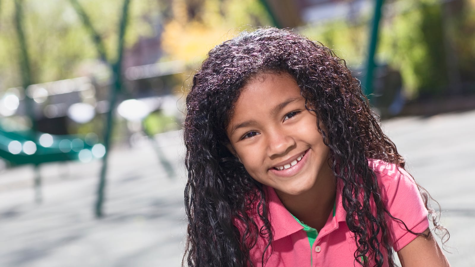 A little girl wearing a pink polo shirt smiles at a playground.